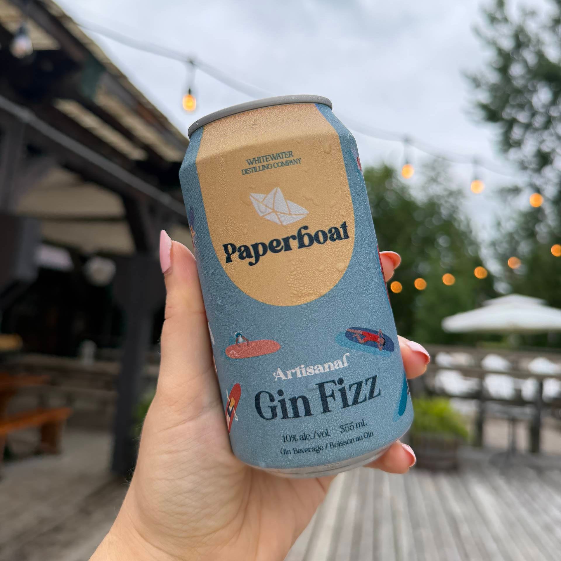 A hand holding a can of Paperboat Artisanal Gin Fizz against an outdoor patio background with string lights and wooden decking.