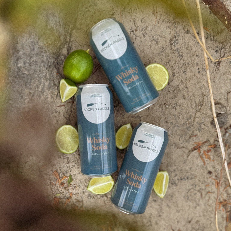Three cans of Broken Paddle Whisky Soda are arranged on a textured stone surface with lime slices around them.
